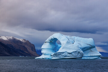 Wall Mural - dark and stormy skies over a blue iceberg that has been carved into an arch by the arctic winds. kai