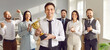 © Studio Romantic - Portrait of a happy smiling young man winner holding the trophy award for success in business standing in office with a team of people colleagues applauding for company best employee reward. Banner