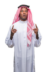 Wall Mural - Young arabic african man wearing traditional keffiyeh over isolated background amazed and surprised looking up and pointing with fingers and raised arms.