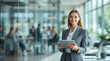 © CreativeCloudStock - Professional woman smiling confidently Wearing gray business suit Holding tablet Modern open office space Large windows Colleagues working Clean professional atmosphere Women selective focus