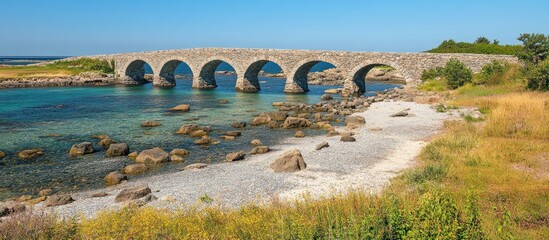 Poster - Stone bridge overlooking the serene sea with clear skies and rocky shoreline surrounded by lush greenery.