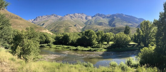 Poster - Scenic landscape with lush greenery and a tranquil river under a clear blue sky showcasing beautiful weather and mountain backdrop
