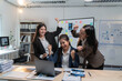 © Tj - Three asian businesswomen are cheering and raising their arms in celebration of a successful business achievement in a modern office