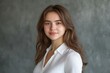 © SerPak - A young woman with long brown hair stands confidently, wearing a white blouse. Her natural expression and soft features create a warm and inviting atmosphere against the smooth gray backdrop.