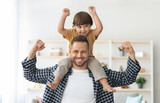 Family power. Strong young man carrying his little son on shoulders, both demonstrating biceps and smiling to camera at home, sport lovers concept