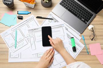 Leinwandbilder - Website designer using smartphone at wooden table with wireframes, stationery and laptop, top view