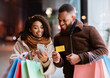 © Prostock-studio - Easy Payment Concept. Portrait of happy african american man and woman using mobile phone, holding credit card and shopping bags, standing outdoors in the evening. Retail And Purchase