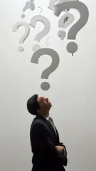 Poster - Businessman with arms crossed is looking up at hanging question marks on a gray background, symbolizing confusion and uncertainty