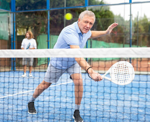 Wall Mural - Concentrated elderly woman padel player hitting ball with a racket on a hard court