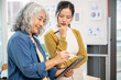© Kittipong - A young Asian female employee is discussing work with her senior female boss in the office, looking at a tablet screen while standing, planning a project together. Startup business, marketing