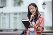 © crizzystudio - .Asian female student smiles happily Standing with a book outside on campus. College students work on educational campuses