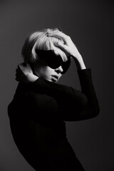 Poster - Stylish woman with short blonde hair in black outfit and sunglasses, posing dramatically against a dark grey background. Fashion and confidence concept.