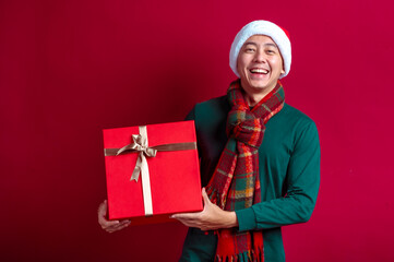 An Asian man wearing a Santa hat, green sweater, and red plaid scarf, holding a festive red gift box with a gold ribbon, standing against a vibrant red background, expressing holiday cheer