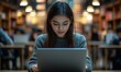© Onchira - Young woman student study in the school library with blurred other people background. She using laptop and learning online
