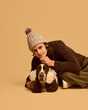 © master1305 - Portrait of young man in warm, cozy chocolate sweater posing with his adorable calm dog wearing warm headwear, against beige studio background. Concept of animal, winter season, friendship, happiness