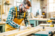© isavira - A carpenter in work uniform working with boards in a carpentry shop surrounded by various tools