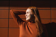 © VISTA by Westend61 - Happy redhead woman in front of brown wall on sunny day