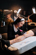 © Xeniya - Charming girl tattoo on a man's leg in a tattoo parlor, creative small business and tattoo master. Hipster woman in gloves draws a tattoo