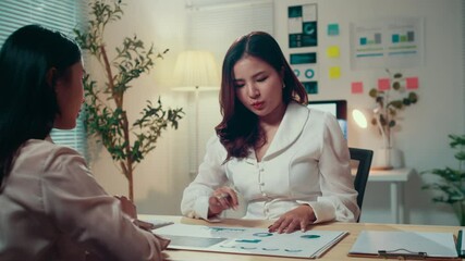 Fototapete - Two businesswomen discuss financial charts at a desk in an office at night, with one using a calculator to analyze data and strategize for future investments