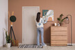 © Pixel-Shot - Young woman hanging painting on brown wall in hall, back view