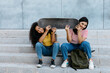 © Tania Cervian/Stocksy - Smiling girlfriends with skateboard sitting on stairs