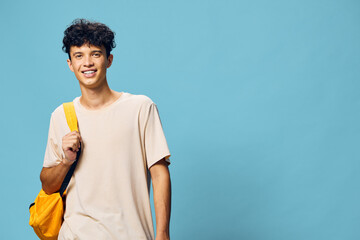 Wall Mural - Young man smiling against a light blue background, wearing a casual beige t shirt and carrying a yellow backpack, embodying a sense of youth and adventure in urban style