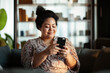 © Olena - Portrait of a happy mixed-race woman using her phone at home. A married woman sitting on the couch, smiling and looking at her phone, engaging in online shopping.