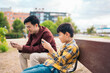 © Santi Nuñez/Stocksy - Father and son sitting on a bench playing video game on phone