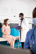 © DC Studio - Doctor discusses prenatal care with pregnant patient while nurse reviews medical records from her desk. Female healthcare worker examines files on laptop as physician gives advice to expecting woman.