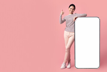 Wall Mural - Cheerful Young Korean Lady Leaning At Big Blank Smartphone And Showing Thumb Up, Smiling Asian Woman Recommending New Mobile App Or Website, Standing Over Pink Studio Background, Mockup Image