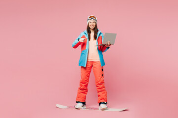 Poster - Full body young IT woman wear warm blue padded jacket ski goggles mask spend weekend winter season in mountains hold use point on laptop pc computer ride snowboard isolated on plain pink background.