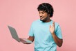 © ViDi Studio - Young IT Indian man he wears blue t-shirt casual clothes hold use work on laptop pc computer waving hand talk speak isolated on plain pastel light pink background studio portrait. Lifestyle concept.