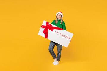 Wall Mural - Full body merry young woman wears green sweater Santa hat posing hold big gift coupon voucher card for store isolated on plain yellow background. Happy New Year celebration Christmas holiday concept.