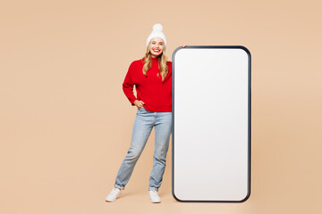 Poster - Full body merry young woman wear red warm cozy sweater hat posing big huge blank screen area mobile cell phone smartphone isolated on plain beige background. Happy New Year Christmas holiday concept.