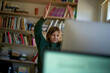 © Connect Images - Smiling child with arms raised in a victory gesture in front of a computer at a desk with a bookshelf in the background.