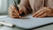 © walaiporn - A hand holding a pen writes on a sheet of paper, reflecting focus and attention to detail in a workspace setting.