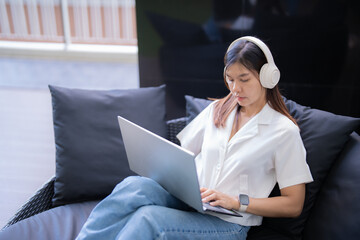 Wall Mural - Young asian woman wearing headphones using laptop while sitting on comfortable sofa and working remotely at home, demonstrating work-life balance and enjoying the freedom of freelance work