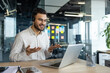 © Liubomir - A young businessman is seen participating in a virtual meeting via his laptop, using a headset in a modern office setting. His expressive gestures reflect active communication and professionalism.
