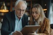 © SerPak - In a warm and inviting library, a young woman and an older man engage in a learning moment. She demonstrates how to use a tablet while he attentively observes, showcasing a meaningful intergenerationa