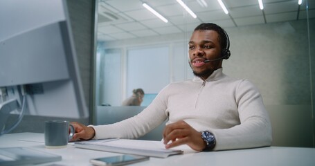 Wall Mural - African American marketing specialist or E-commerce manager talks on headset with client, uses computer, provides online customer service. Young businessman works in bright modern office. E-business.