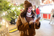 © Home-stock - Happy young lady using smartphone in the city with festive Christmas fair background, texting online while walking on winter holidays