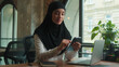 © Yuliia - Muslim woman in hijab islamic girl Arabian businesswoman entrepreneur calm smiling business lady working at desk scrolling mobile phone gadget addicted female online app cellphone internet in office