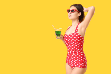 Wall Mural - Beautiful young Asian woman in swimsuit with glass of cocktail on yellow background