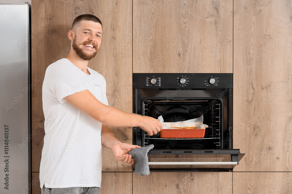 Young man taking baking dish with potato from oven in kitchen