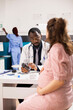 © DC Studio - Physician explains importance of prenatal vitamins to pregnant white woman in medical office while in background african american nurse is working. Doctor gives expectant lady pregnancy medication.