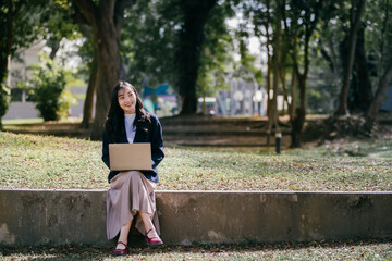 Aufkleber - Young businesswoman sitting on a low wall in a park, working on her laptop while soaking up the fresh air and warm sunshine, blending productivity with outdoor tranquility
