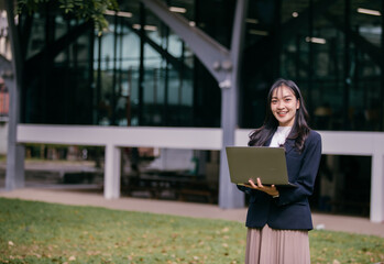 Aufkleber - Young asian businesswoman is holding a laptop and smiling in a university campus park, enjoying the green space and fresh air while working remotely