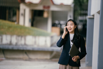 Poster - Young businesswoman smiling while engaging in a phone conversation, holding a laptop and enjoying her workday outdoors in a vibrant urban setting, exuding confidence and professionalism