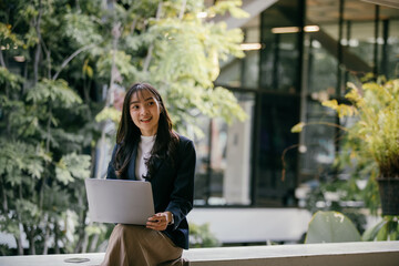 Aufkleber - Young businesswoman is sitting on a low wall outside a modern office building, smiling as she works on her laptop, enjoying the fresh air and greenery surrounding her