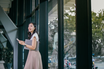 Poster - Smiling businesswoman using her phone and carrying her laptop while walking near the large windows of a modern office building, enjoying work-life balance